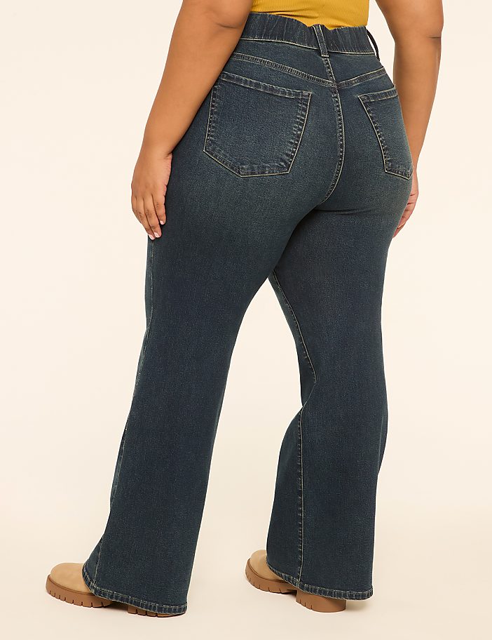 A person wearing dark wash flare jeans and tan shoes stands against a plain, light-colored background.
