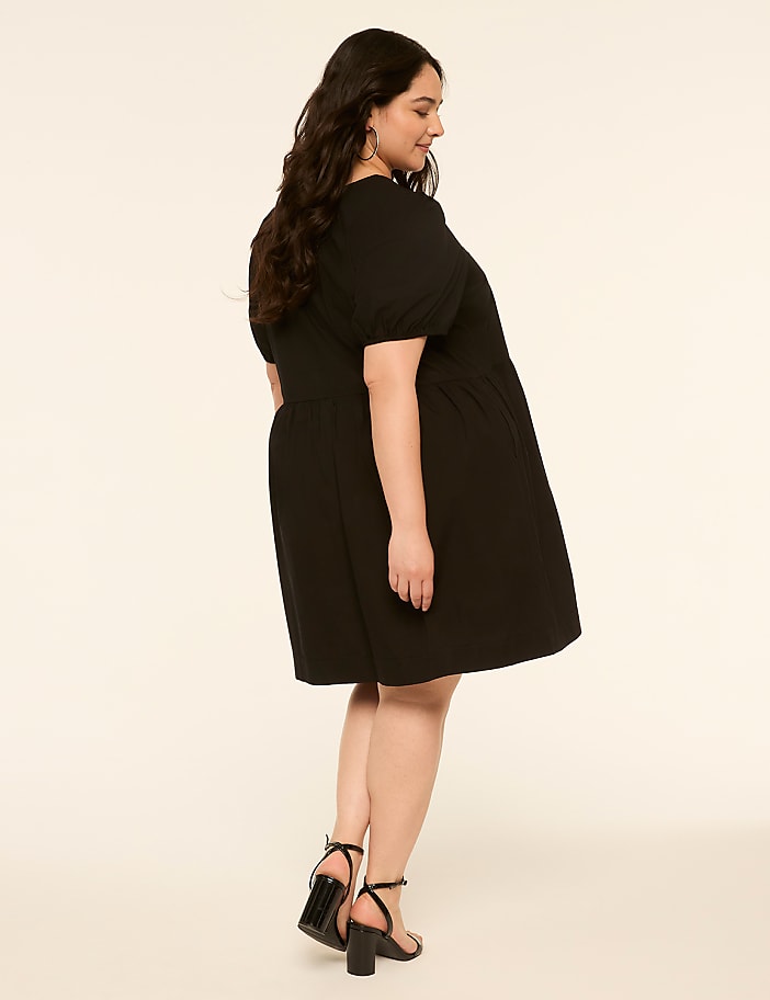 A woman in a black dress and heels stands against a plain, light-colored background.