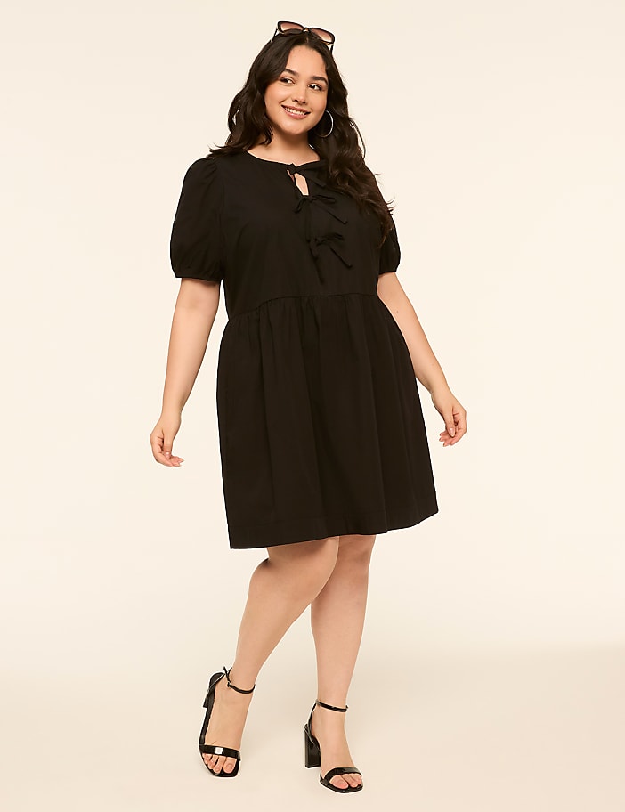 A smiling woman in a black dress stands against a plain, light-colored background.