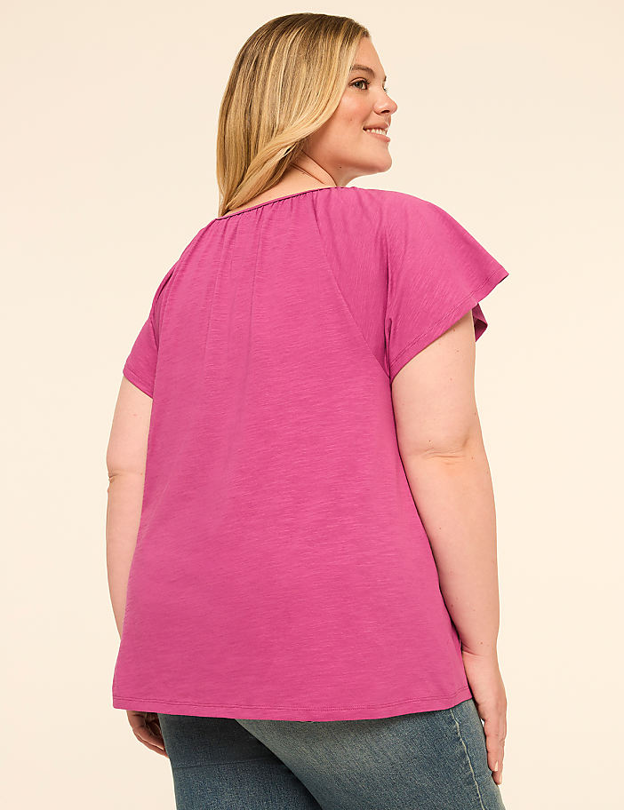 A woman in a pink top and blue jeans stands against a plain, light-colored background.