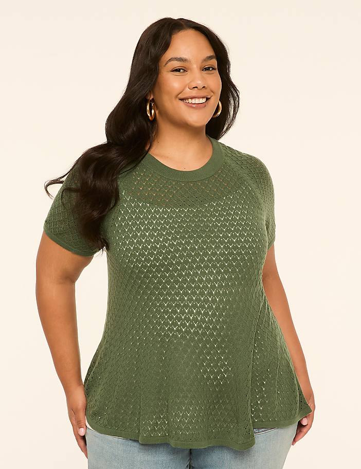 A smiling woman with dark hair and gold hoop earrings wears a green, short-sleeved, textured top and blue jeans against a plain, light-colored background.