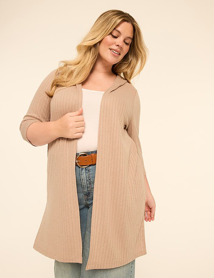 A woman in a beige hooded cardigan and jeans stands against a plain, light-colored background.