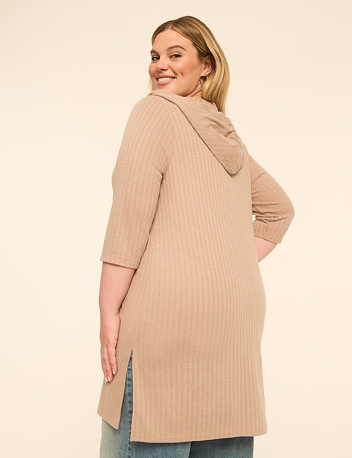 A woman in a beige hooded tunic and jeans smiles over her shoulder against a plain, light-colored background.