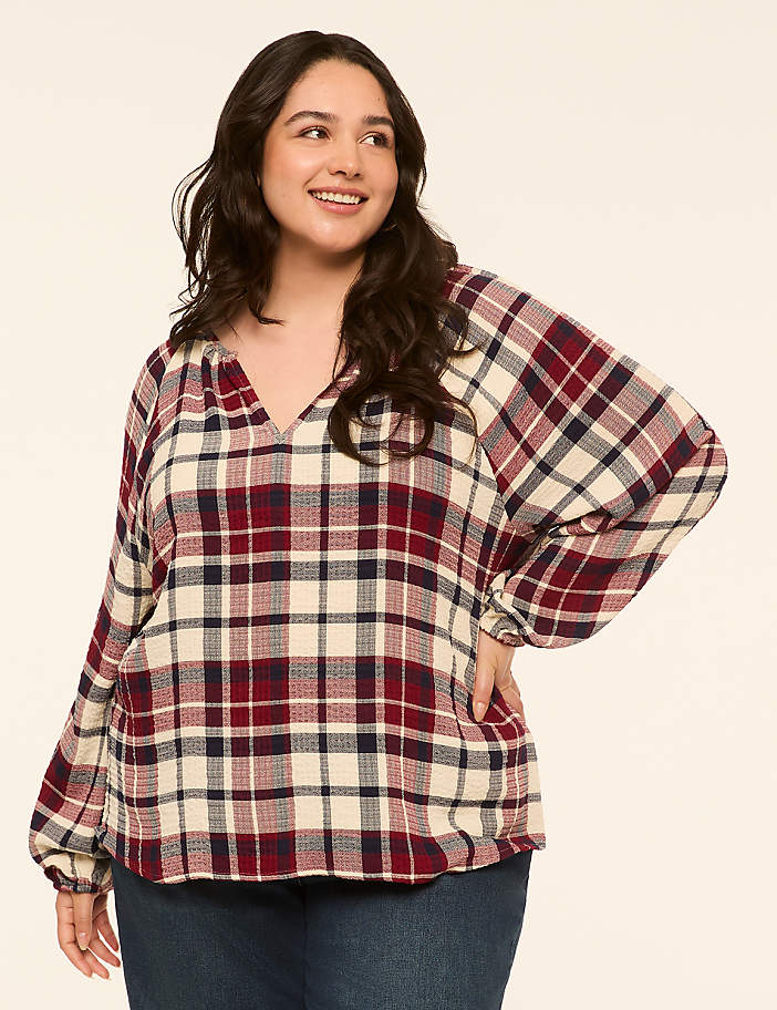 A smiling woman in a plaid blouse and jeans stands against a plain, light-colored background.