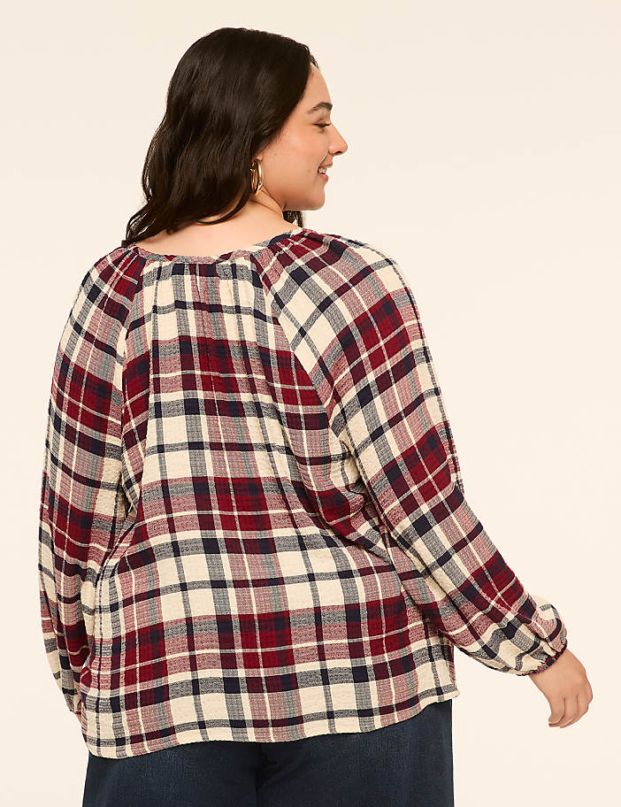 A woman in a red, white, and blue plaid shirt stands against a plain, light-colored background.