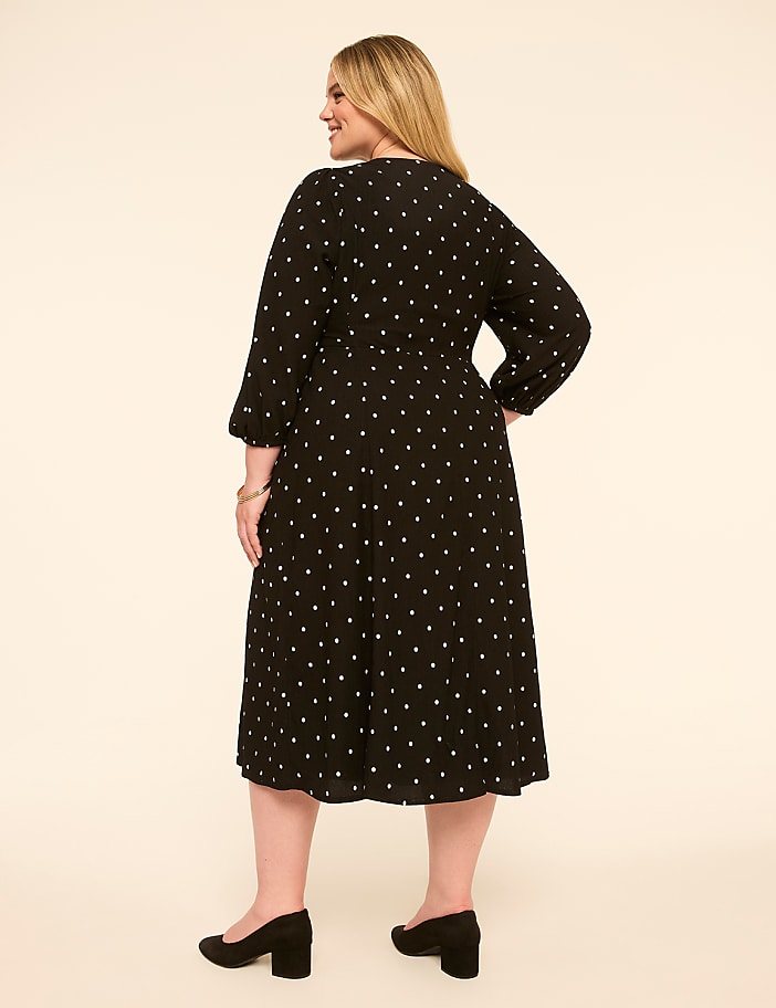 A woman in a black polka dot dress stands against a plain, light-colored background.