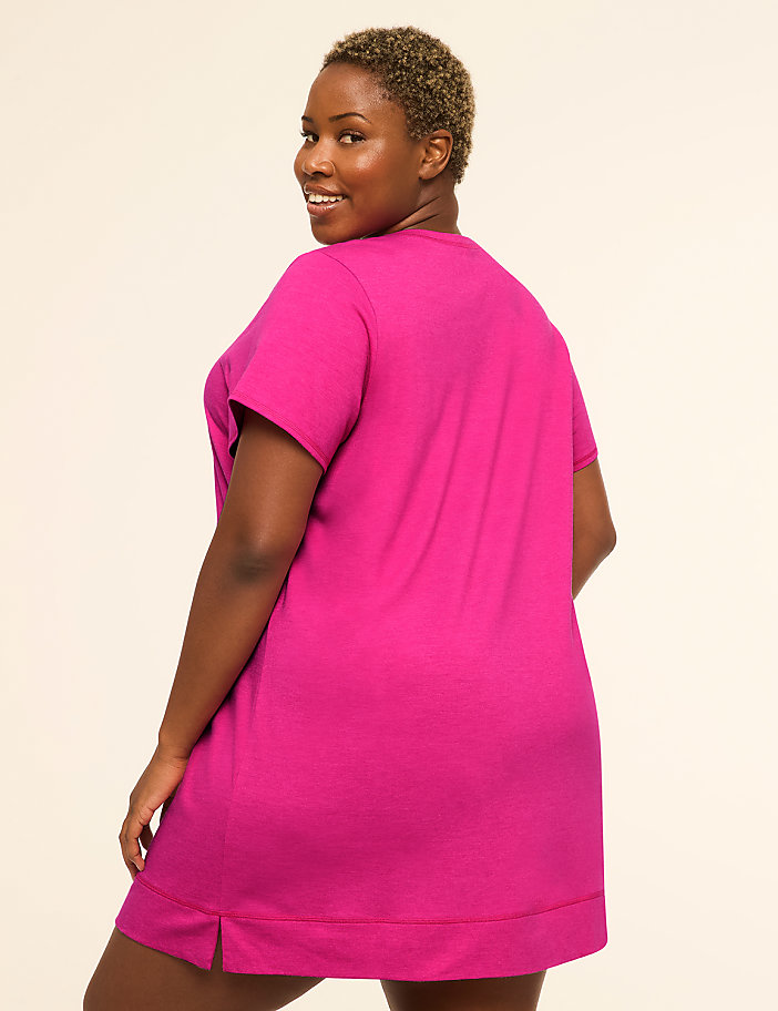 A woman in a bright pink t-shirt dress smiles over her shoulder against a plain, light-colored background.