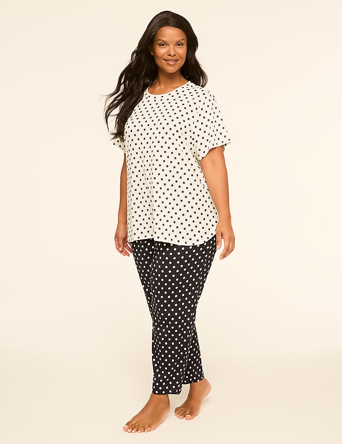 A woman with long dark hair stands in the foreground wearing a white polka dot shirt and black polka dot pants against a plain, light-colored background.