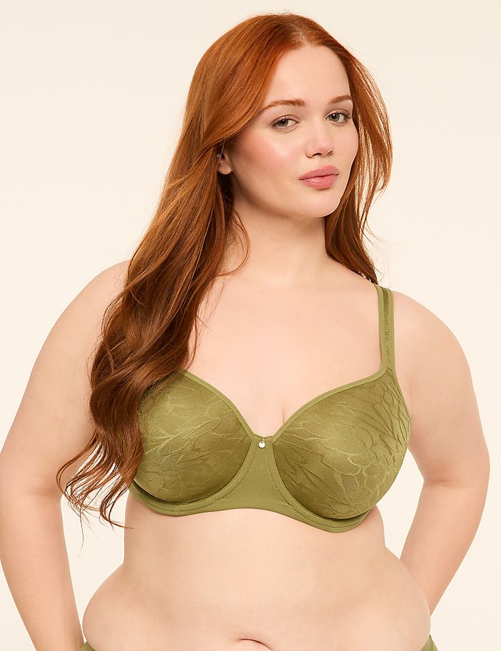 A woman with long red hair wears a green textured bra against a plain, light-colored background.