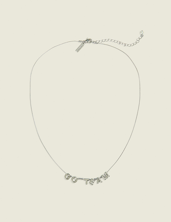 A delicate silver necklace with "GO TEAM" spelled out in block letters rests on a plain, light beige background.