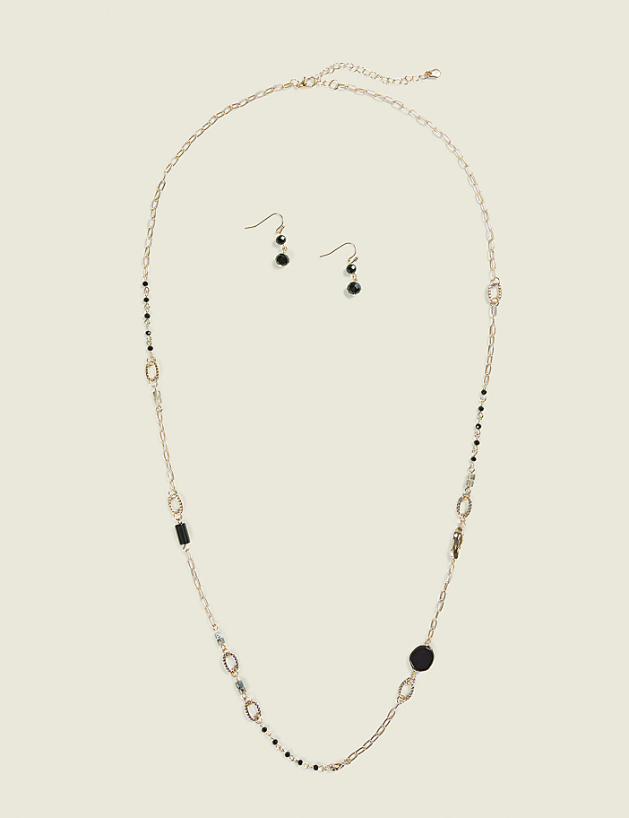 A long gold necklace with black and silver beads and charms, along with a pair of matching earrings, are laid out on a plain, light beige background.
