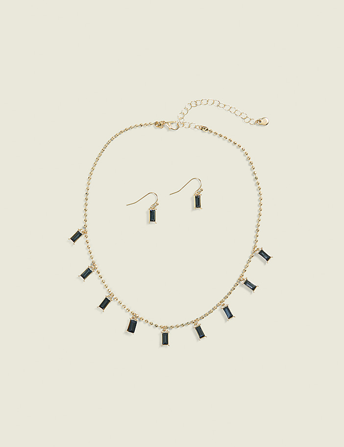 A gold necklace with dangling rectangular blue gems and matching earrings rests on a plain, light beige background.