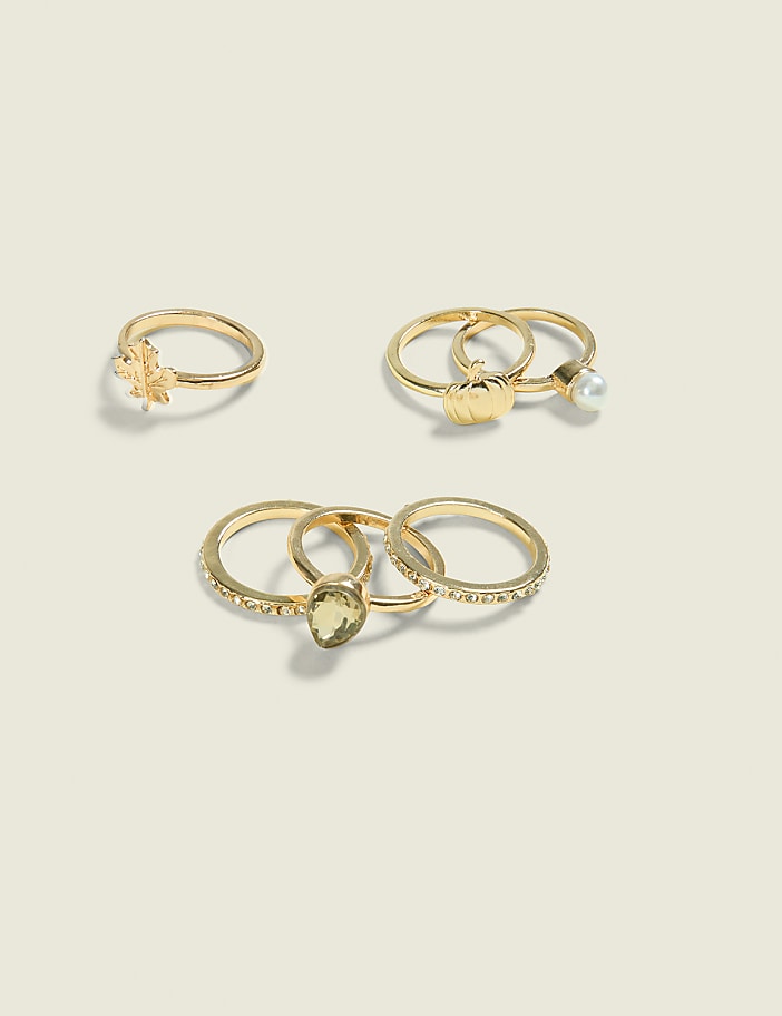 A collection of gold rings, including one with a leaf design, a pumpkin and pearl ring, and a set with a green stone and rhinestones, are arranged on a pale, textured background.