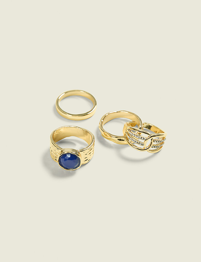Four gold rings, one with a blue stone and another with small diamonds, are arranged on a plain, light-colored background.