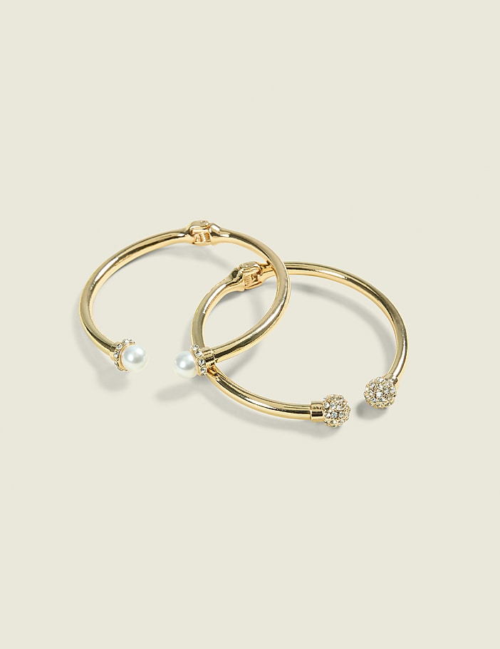 Two gold bangles, one adorned with pearls and the other with rhinestones, rest on a plain, light-colored background.