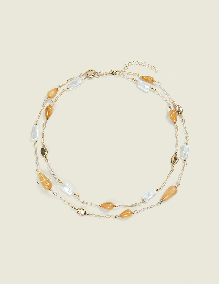 A delicate gold chain necklace with irregular white pearls and amber-colored beads is laid out on a plain, light beige background.