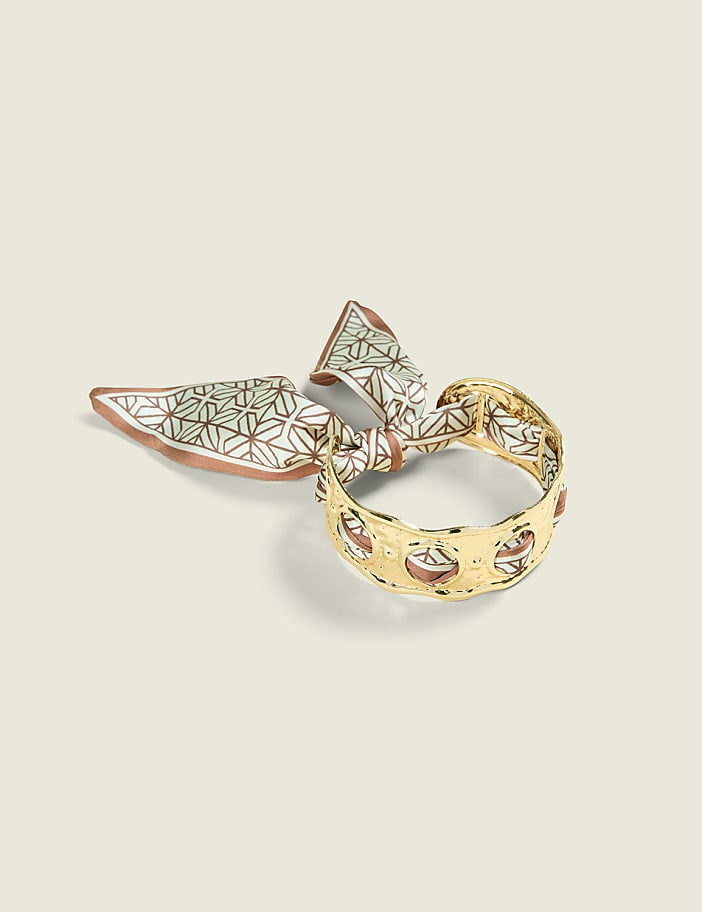 A gold bracelet with a patterned silk scarf tied around it rests on a pale, textured background.