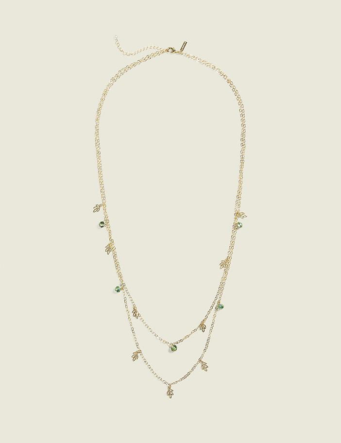 A delicate gold layered necklace with small green gemstone charms hangs against a plain, light beige background.