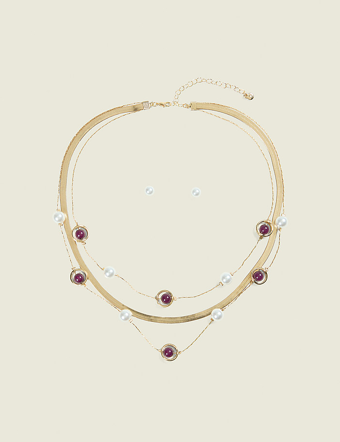 A layered gold necklace with pearls and purple beads rests on a plain, light-colored background, with a pair of pearl stud earrings positioned above it.