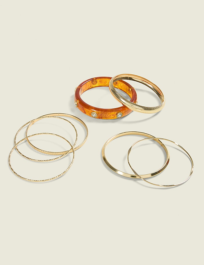 Several gold bangles and a decorative orange bangle with rhinestones are arranged on a plain, light-colored background.
