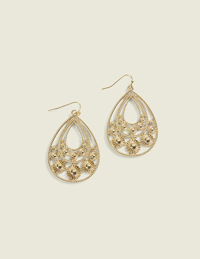 A pair of ornate gold teardrop earrings with intricate floral designs hang against a plain, light beige background.