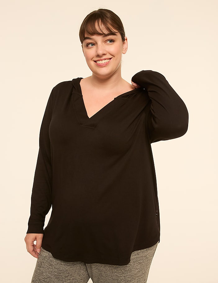A smiling woman in a black hooded shirt and gray sweatpants stands against a plain, light-colored background.