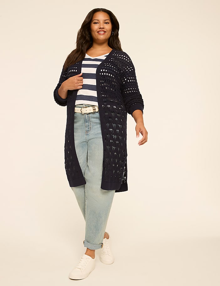 A woman in a striped shirt, jeans, and a long navy cardigan stands against a plain, light-colored background.