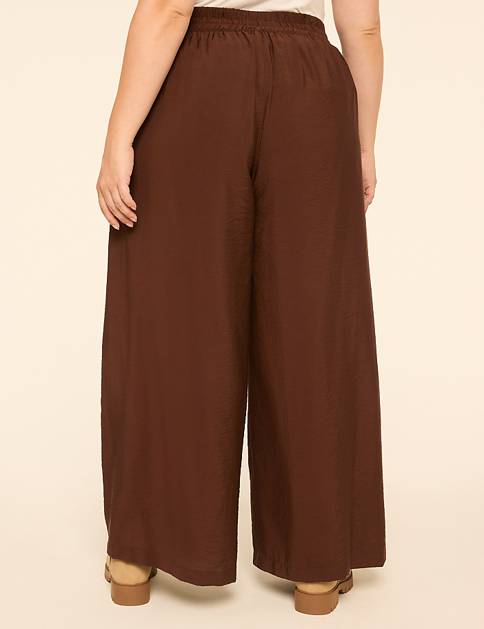 A person wearing brown wide-leg pants and sandals stands against a plain, light-colored background.