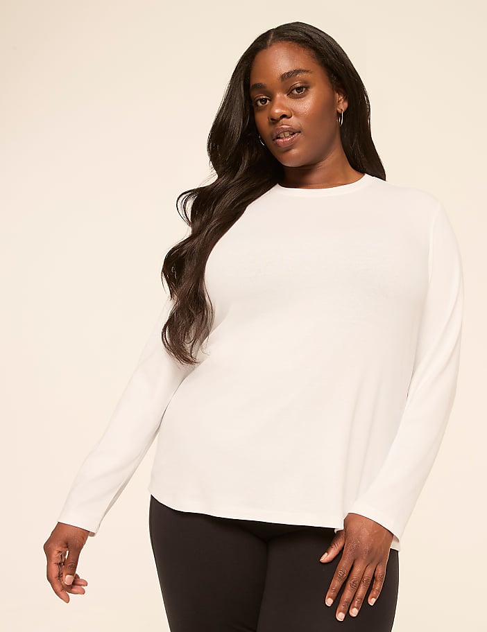 A woman with long, dark hair wears a white long-sleeved shirt and black leggings against a plain, light-colored background.