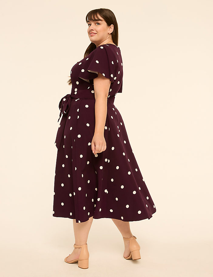 A woman in a polka dot dress stands against a plain, light-colored background.