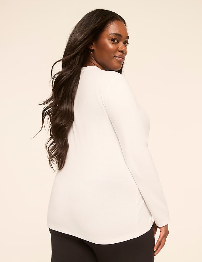 A woman with long, dark hair wearing a white long-sleeved shirt and black pants turns her head to smile over her shoulder against a plain, light-colored background.
