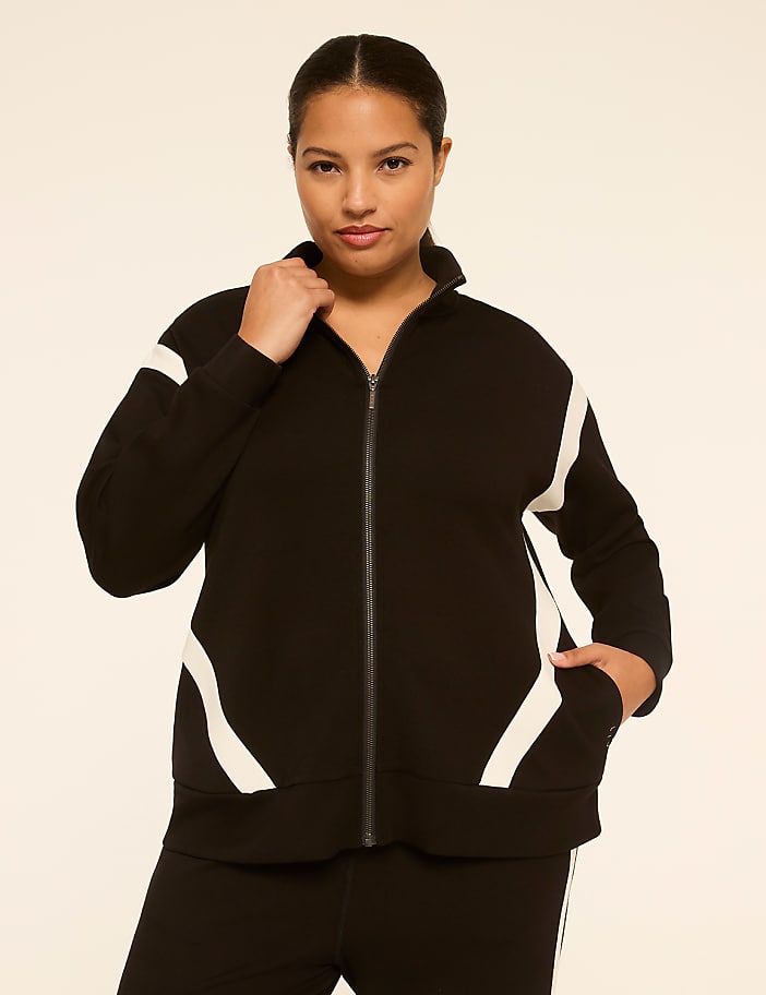 A woman in a black zip-up jacket with white accents stands against a plain, light-colored background.