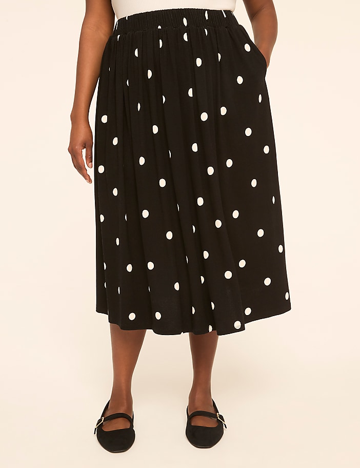 A person wearing a black polka dot skirt and black flats stands against a plain, light-colored background.