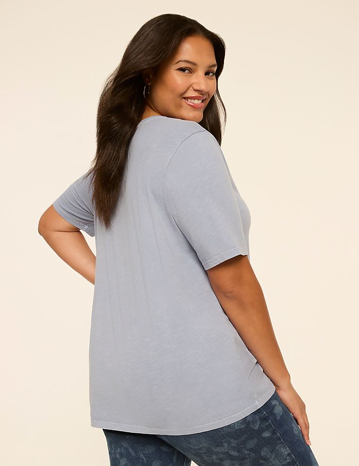 A woman in a light blue t-shirt and patterned jeans smiles over her shoulder against a plain, light background.