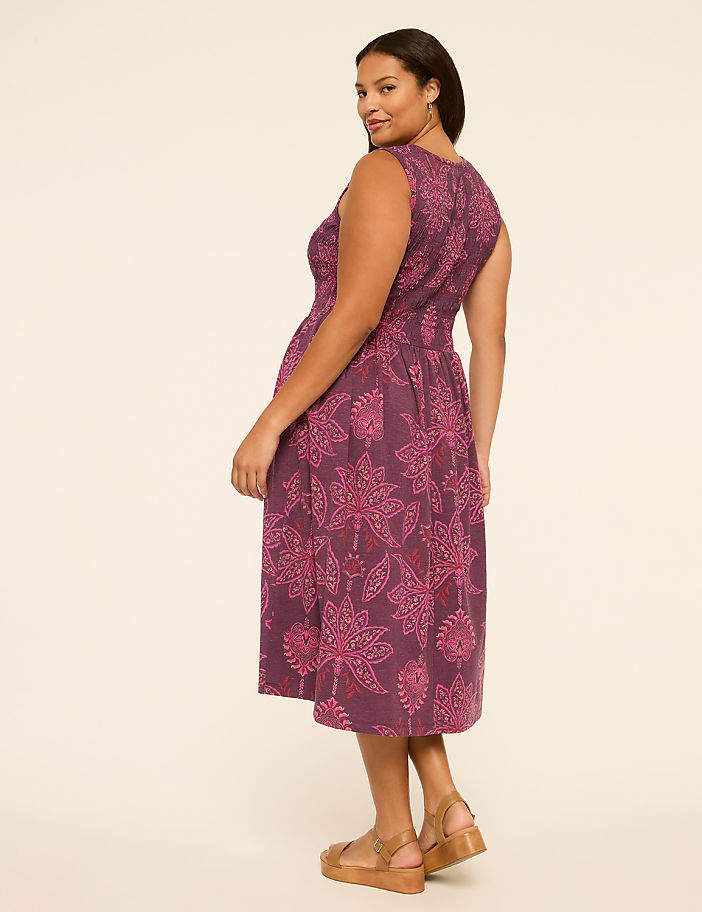 A woman in a floral purple dress stands against a plain, light-colored background.