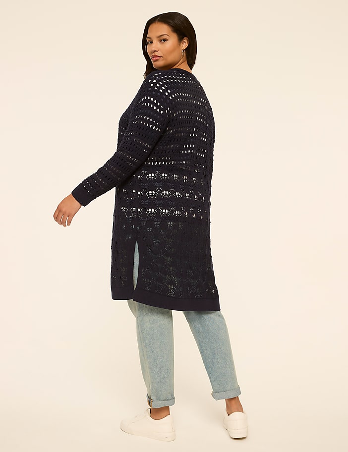 A woman in a navy crocheted cardigan and light wash jeans stands against a plain cream background.