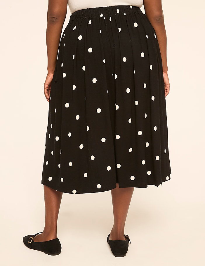 A person wearing a black polka dot skirt and black flats stands against a plain beige background.
