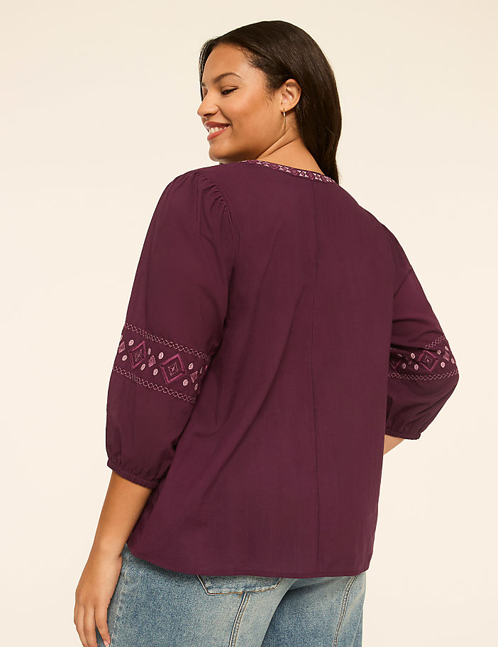 A woman in a purple embroidered blouse and jeans stands against a plain, light-colored background.