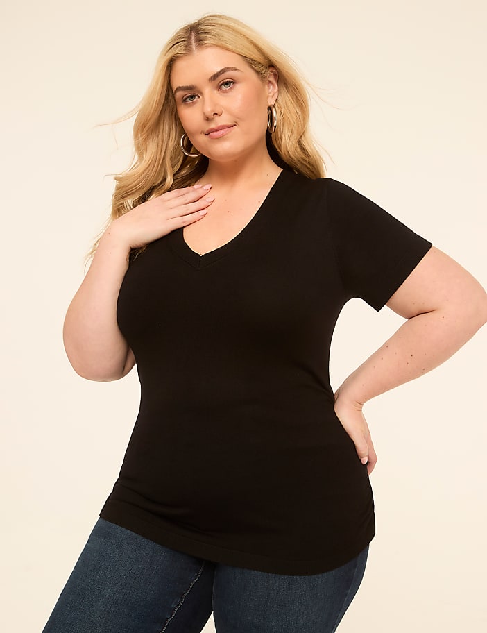 A woman in a black v-neck shirt and blue jeans poses against a plain, light-colored background.
