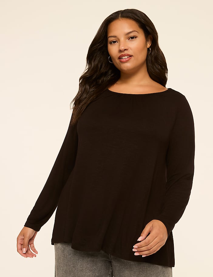 A woman in a black long-sleeved shirt and gray jeans stands against a plain, light-colored background.