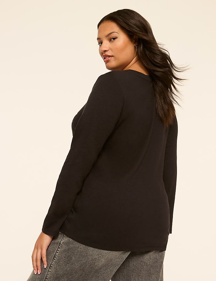 A woman in a black long-sleeved shirt and gray jeans stands against a plain, light-colored background.