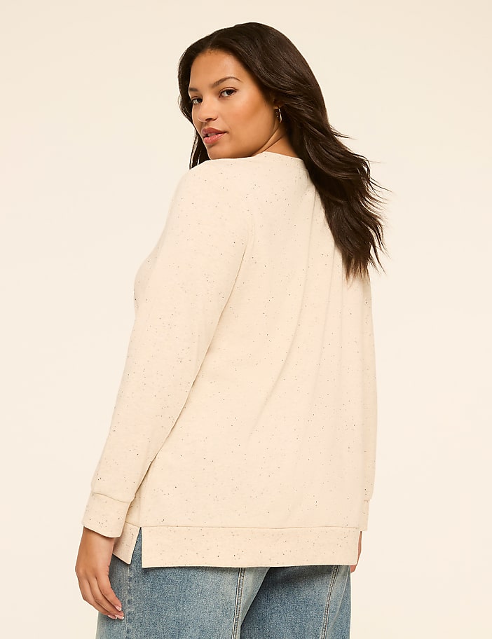 A woman in a cream-colored sweater and blue jeans stands against a plain, light-colored background.