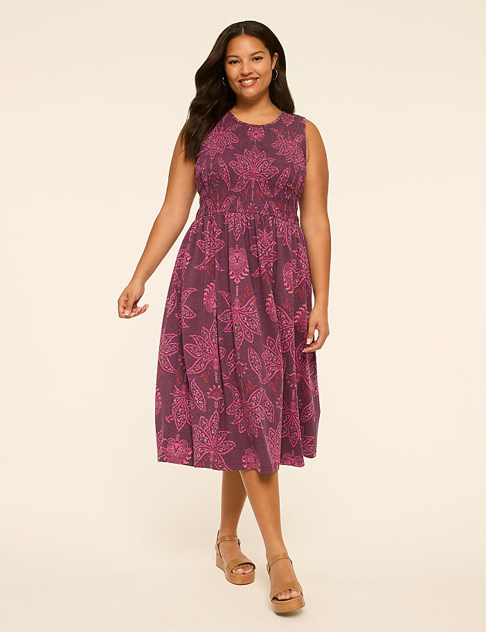 A smiling woman in a purple floral dress stands against a plain, light-colored background.