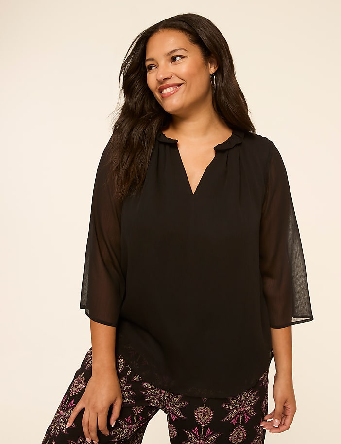A smiling woman with dark hair wears a black sheer blouse and patterned pants against a plain, light background.