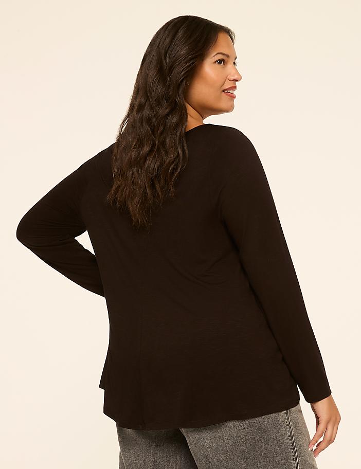 A woman in a black long-sleeved shirt and grey jeans stands against a plain, light brown background.