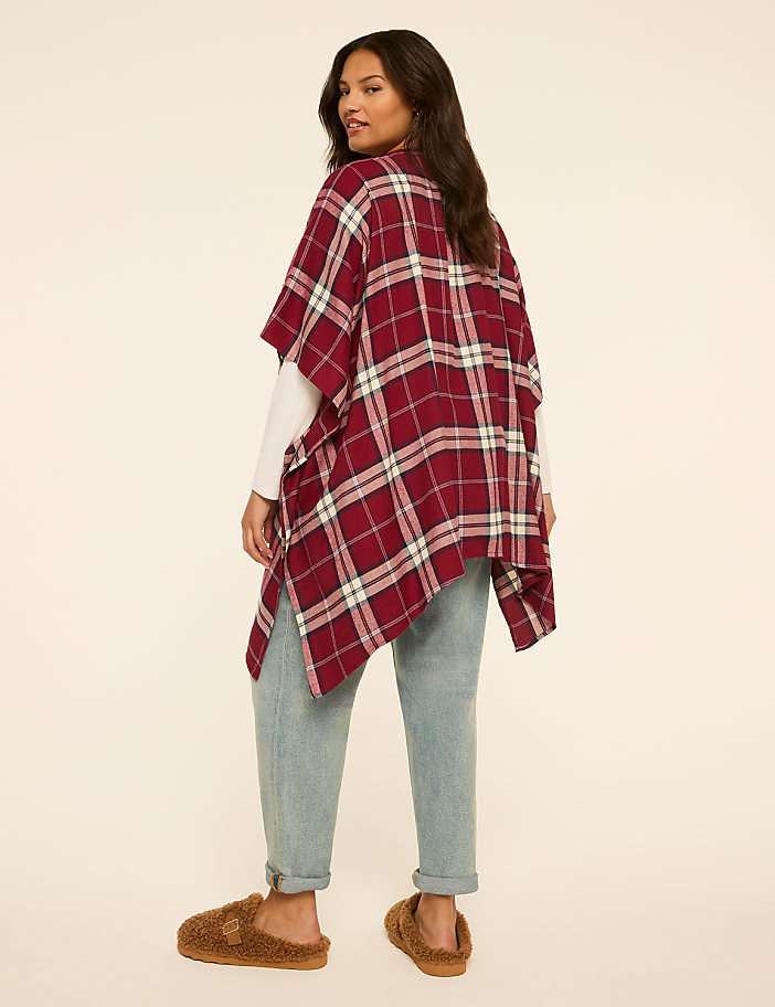 A woman in a plaid poncho and jeans stands on a plain background.