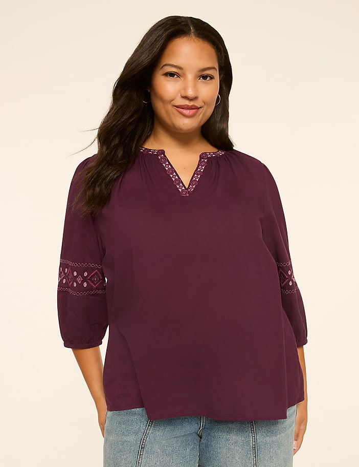 A woman in a plum-colored embroidered top and jeans stands against a plain, light-colored background.