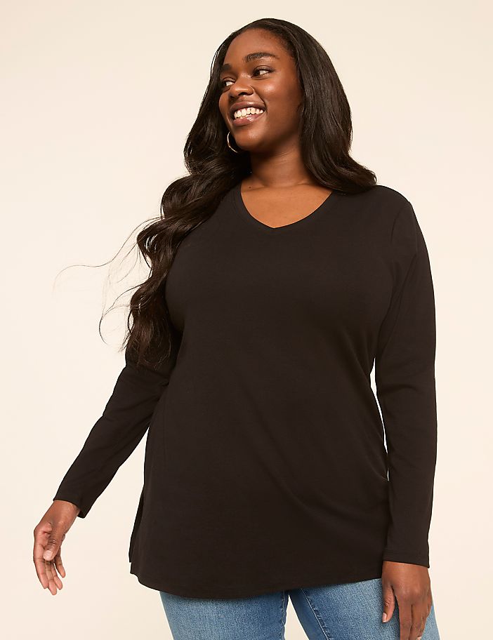 A smiling woman with long dark hair wears a black v-neck long-sleeved shirt and blue jeans against a plain, light-colored background.