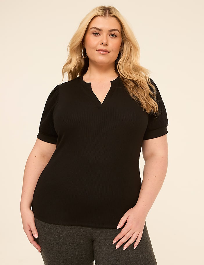 A woman with blonde hair and a black top stands against a plain, light-colored background.