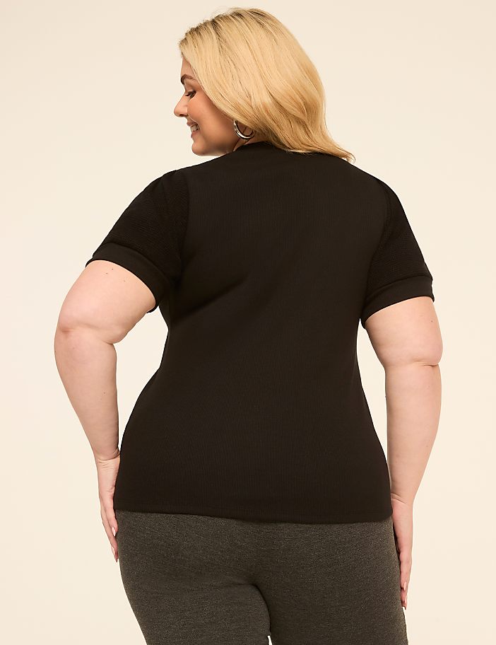A woman in a black ribbed top and grey pants stands against a plain, light-colored background.